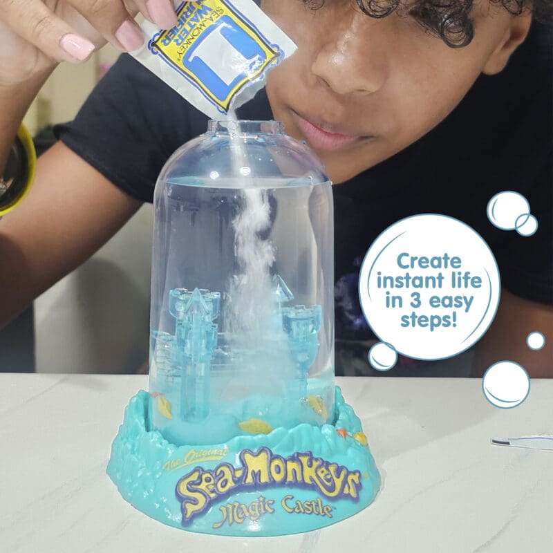 23230-Sea-Monkeys Magic Castle-Image3 A child wearing a dark shirt and pink nailpolish is bent over the aquarium, and pouring powder into it from a small packet labeled with the number 1. There is a callout in a circle that reads "Create instant life in 3 easy steps!".